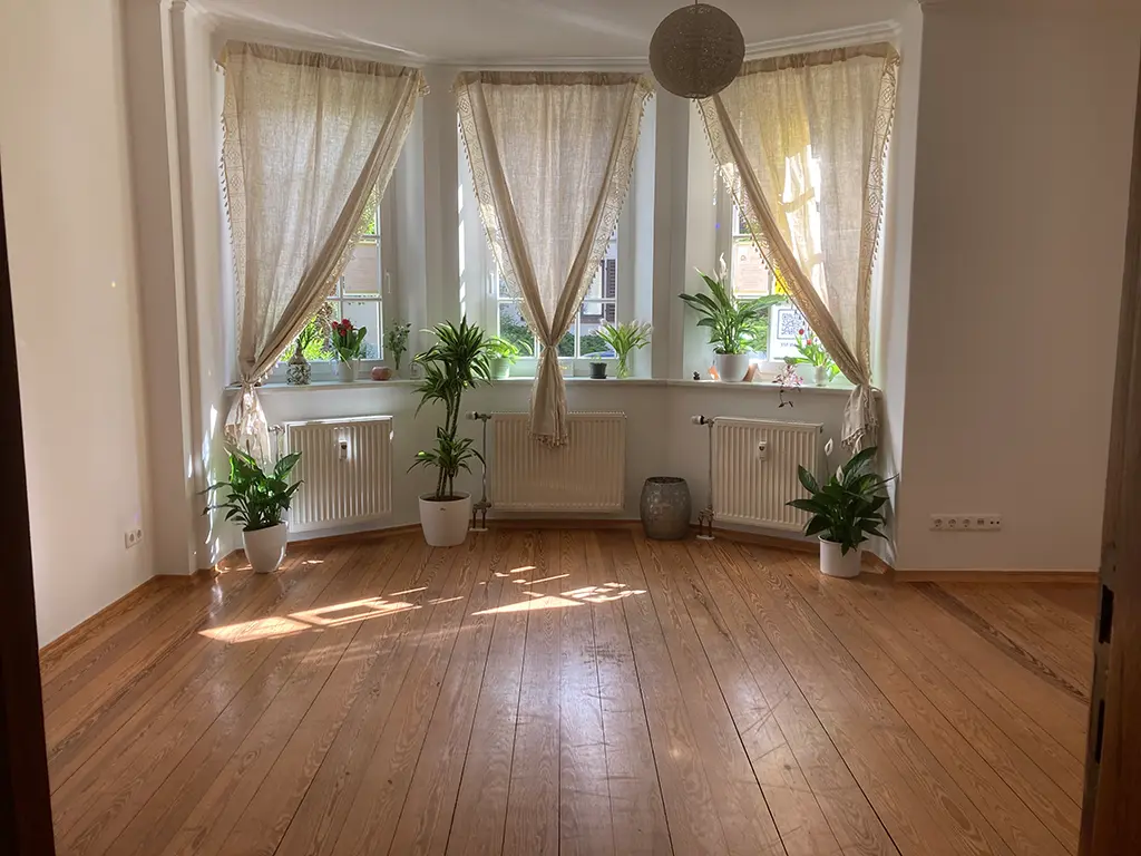 Heller Raum mit Holzboden, drei Fenstern mit beigen Vorhängen, Zimmerpflanzen und Sonnenlicht auf dem Boden.