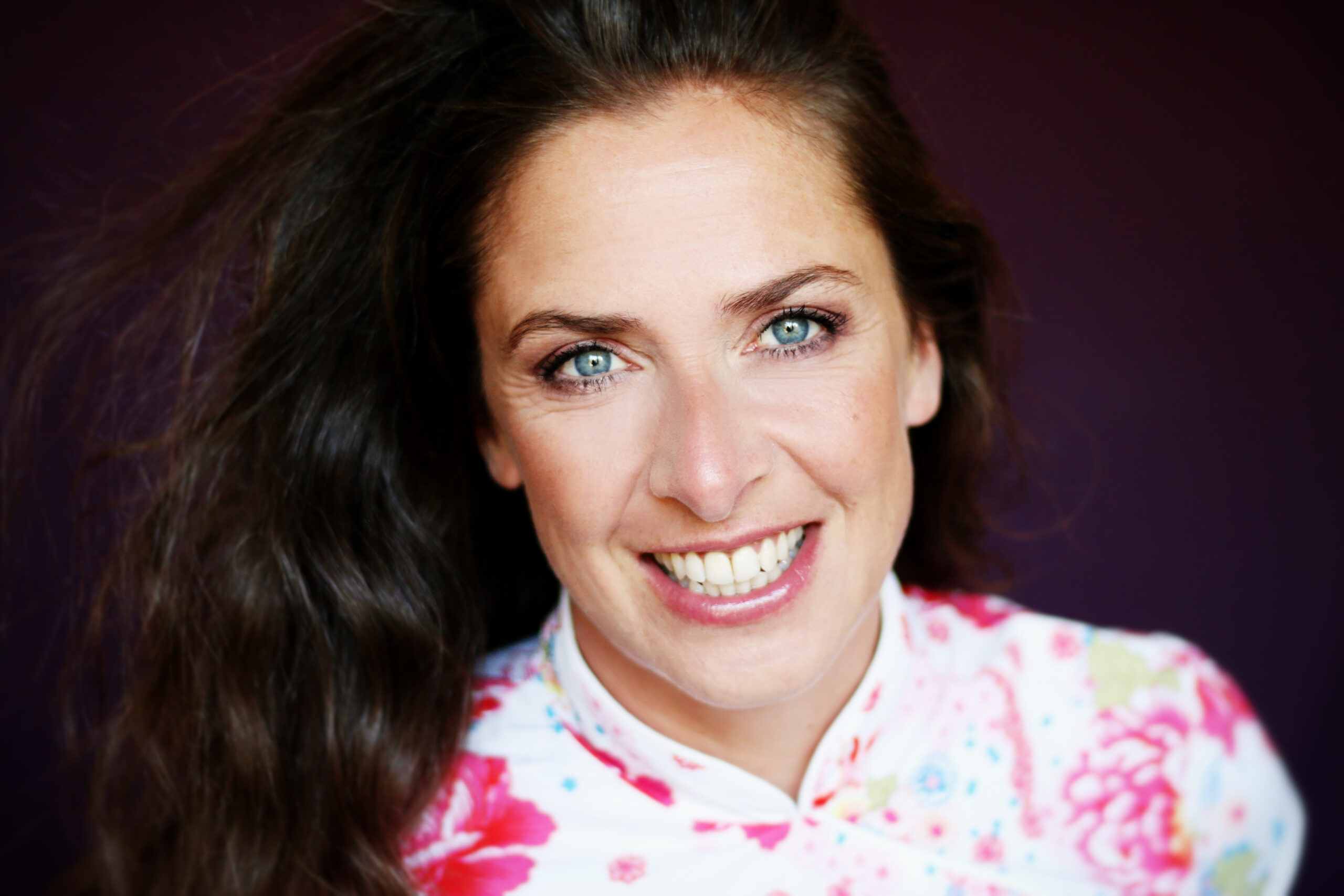 Porträt einer lächelnden Frau mit langen dunklen Haaren und hellen Augen vor dunklem Hintergrund. Sie trägt eine helle Bluse mit pinkem Blumenmuster.